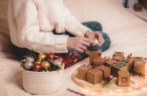 Hands caucasian teenage girl in a white knitted sweater takes out a candy ball from a box of advent calendar while sitting on the bed with christmas decorations in the bedroom, close-up side view.