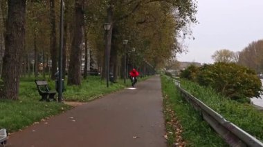 Kırmızı yağmurluk ve miğferli bir adam, bir tarafında alçak kirişler olan bir bisiklete biniyor, diğer tarafında park var, yan görüş yavaş çekimde..