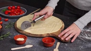 Kafkasyalı genç bir kızın elleri bıçakla kesilmiş, ay şeklinde pizza hamuru şeklinde, koyu çimento bir masada malzemelerle duruyor, yandan yavaş çekimde. Bayram pizzası pişirme kavramını adım adım kavrayın..