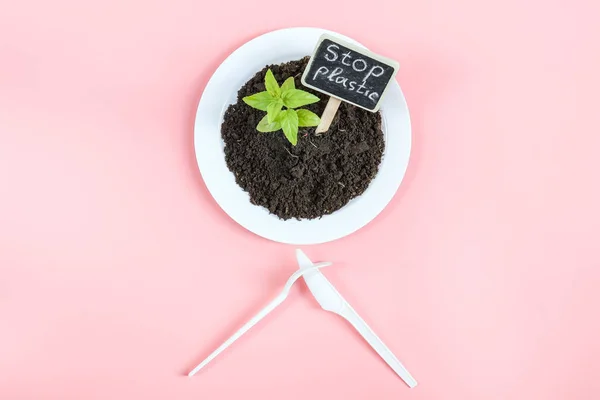 Plastic knife and fork crossed, white plate with black earth, green sprout and peg with the inscription: stop plastic lying on a light pink background, flat lay close-up. The concept of ecology and plastic waste, disposable plastic tableware and rest