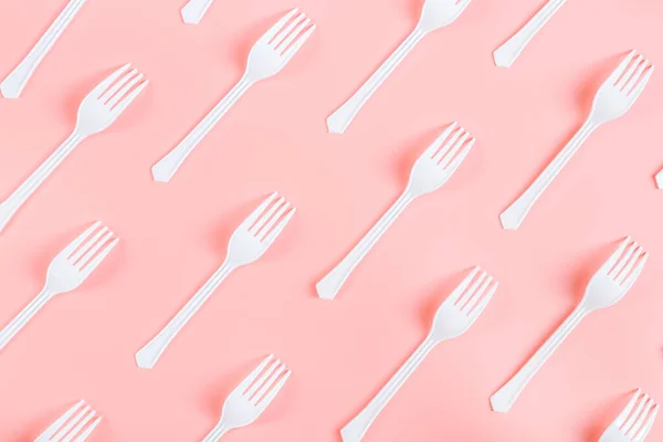 A lot of white plastic forks lie diagonally on a light pink background, flat lay close-up. The concept of ecology, plastic garbage and disposable plastic tableware.