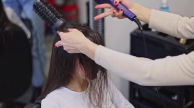 Güzel, beyaz, sarışın kadın kuaför saç kurutma makinesiyle uzun kahverengi saç stili ve fırça başıyla müşteriye doğru eğilmiş saç fırçası kuaförde oturuyor, yan görüş yakın çekim ve yavaş çekim..
