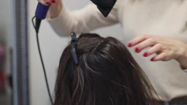 Güzel, beyaz, sarışın kadın kuaför saç kurutma makinesi ve saç köklerini kurutan uzun kahverengi saçlı kuaför kuaförde arka koltukta oturan kadın müşteriye yakın çekim ve yavaş çekim..