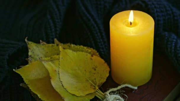 一只黄色的燃着的蜡烛 一束秋天的白杨树叶 还有一本褐色的书 裹着一件深绿色的针织毛衣在床上 在慢镜头中近距离观看 然后放大 — 图库视频影像