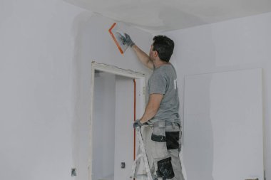Gri üniformalı ve eldivenli yakışıklı beyaz bir adam, merdiven üzerinde duruyor, akşamları kapısında süngerimsi bir malayla yeni sıvanmış bir duvarı düzleştiriyor, bina feneri de açık, yakından bakıyor. İnşaat konsepti.