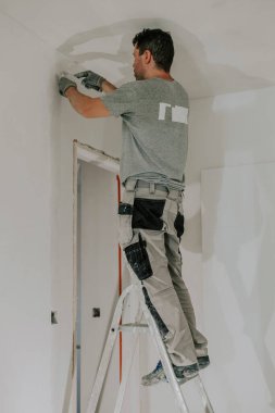 Gri üniformalı yakışıklı bir beyaz adam ve merdiven üzerinde duran eldivenler akşamleyin kapının önündeki duvara iki küçük spatula koyuyor ve bir bina feneri açık, yan görüş açısında. İnşaat konsepti.