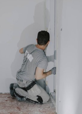 Gri üniformalı ve eldivenli yakışıklı beyaz bir adam, arkadan çömelmiş, küçük bir spatula ile kapı aralığını kokutuyor, yakından bakıyor. İnşaat konsepti.