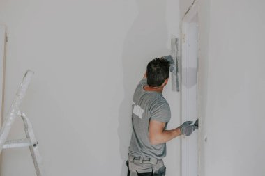 Gri üniformalı yakışıklı beyaz bir adam ve arkada duran eldivenler elinde küçük bir spatula tutuyor ve kapı aralığının geniş bir açısını, yakından bakıyor. İnşaat konsepti.