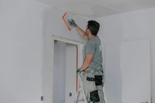 stock image A handsome young Caucasian man in a gray uniform and gloves, standing on a stepladder, smooths a freshly plastered wall in a doorway with a spongy trowel in the evening with a building lantern on, close-up side view. Construction concept.