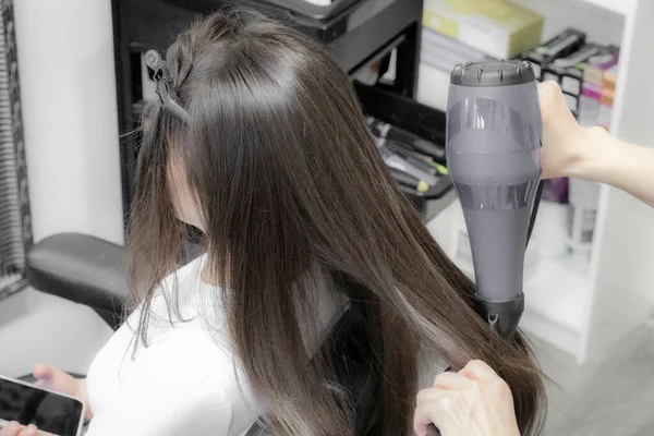 一位年轻的高加索女理发师用吹风机擦干她的头发 把她的一头棕色长发梳在椅子上 坐在美发厅里 侧视特写和有选择的焦点 理发店的概念 — 图库照片