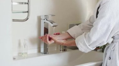 Beyaz bornozlu beyaz bir kadın, sağ elini banyodaki musluktan akan temiz suyun altında ağır çekimde yakın çekimde duruluyor. Konsept vücut bakımı, sağlıklı yaşam tarzı.