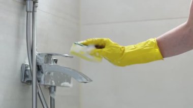 The hands of a caucasian young woman in yellow gloves rub a dirty faucet in the bathroom with a sponge and detergent, side view close-up in slow motion. Cleaning the bathroom concept.