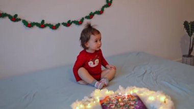 Kafasında at kuyruğu ve noel kıyafetleri olan güzel beyaz bir kız bebek yatakta dans eder ve gelen takvimden çikolatalı tatlılar yedikten sonra şarkı söyler. Yavaş çekimde yan çekimde..