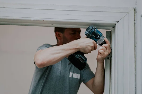 stock image One handsome young male builder stands in a doorway and unscrews the screws of the sling with a drill, close-up side view. Concept of construction work.