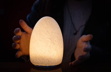 Soyut ve desenli bir yüzeyi olan yumurta şeklindeki bir gece lambası sarı ve sıcak bir ışıkla aydınlatılır ve genç bir kızın elleri onu kavramaya çalışır..