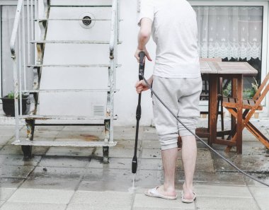 Gri tişörtlü ve arkadan eşofmanlı beyaz bir adam evinin arka bahçesindeki terası yıkıyor. Karcher marka, yan manzaralı, yakın plan. Güneş pansiyonları, ev temizliği.