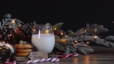 Beyaz örgü kazaklı beyaz bir kadının eli Noel Baba için sütle birlikte bir bardağa pipet koyar. Kurabiye, koni ve ahşap bir masanın üzerinde yanan bir çelenk ile birlikte..