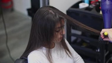 Güzel, beyaz, sarışın kadın kuaför saç kurutma makinesiyle uzun kahverengi saç stili ve saç fırçasıyla kuaförde sandalyede oturan bir müşteriye saç teli çekerek, yakın çekim ve yavaş çekim yaparak ve zoom yaparak..