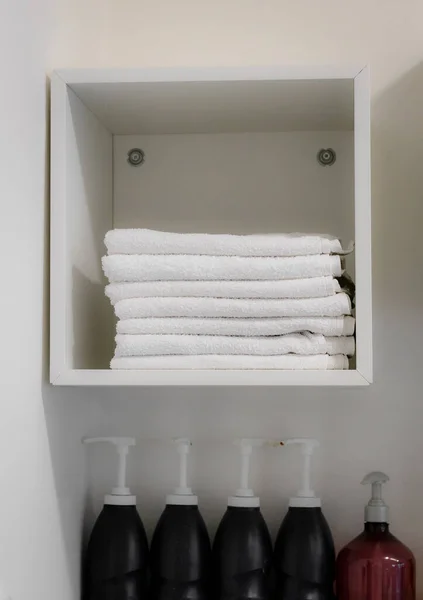 A stack of clean and beautifully shaped terry towels on a white hanging shelf and a row of bottles of shampoos and hair masks with dispensers from below in a hairdressing salon, close-up side view.
