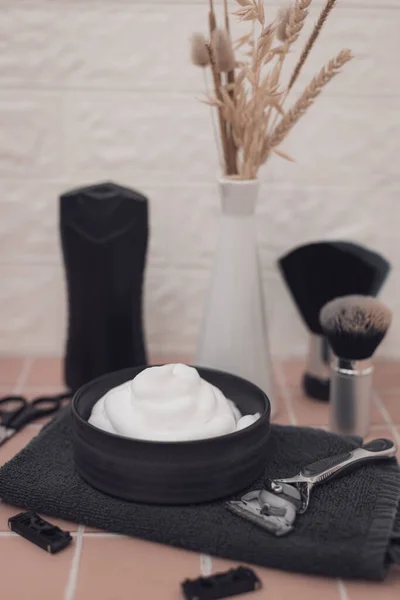 Shaving foam in bowl, shaving brush, razor, razor heads, scissors, bottle of aftershave cream and vase with dried flower on brick tiled background, close-up side view. The concept of a male hairdresser, beauty salon, beard shaving.