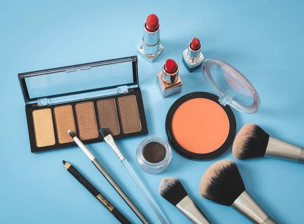 A set of cosmetics from a palette of nude eye shadows, makeup brushes, red lipsticks and an eyebrow pencil on a blue background, close-up top view. The concept of cosmetics, beauty salon.