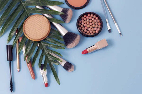 A set of cosmetics from a face powder box, red lipsticks, makeup brushes, mascara and pencils for lips and eyebrows with a palm branch on a blue background, flat lay close-up. The concept of cosmetics, beauty salon.