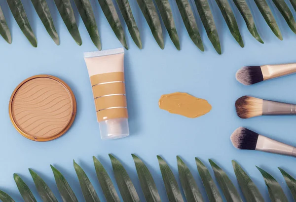 A set of cosmetics from a tube of foundation with a smear, a face powder box and three makeup brushes and a palm branch on a blue background, flat lay close-up. The concept of cosmetics, beauty salon.
