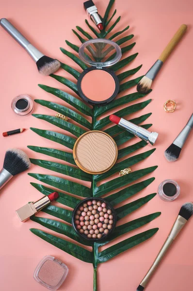 A set of cosmetics from face powder boxes, makeup brushes, red lipstick, eye shadow and rings on a palm tree branch on a pink background, flat lay close-up of sharpness. The concept of cosmetics, beauty salon.