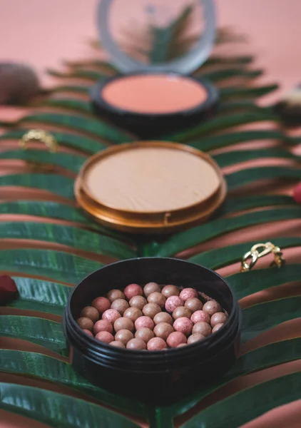 Granular face powder box and gold ring with hard texture powder boxes in blur on a palm branch on a pink background, side view close-up of the field. The concept of cosmetics, beauty salon.