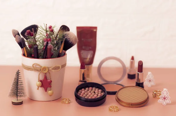 A set of cosmetics from face powder boxes, makeup brushes in a pot, red lipsticks, rings and christmas decorations on a pink background, close-up side view with depth of field. The concept of cosmetics, beauty salon.
