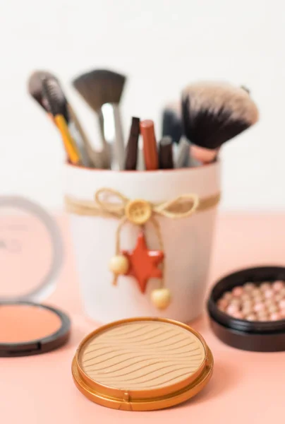 A set of cosmetics from face powder boxes and makeup brushes with eyeliners in a pot with christmas decorations on a pink background, close-up side view with depth of field. The concept of cosmetics, beauty salon.