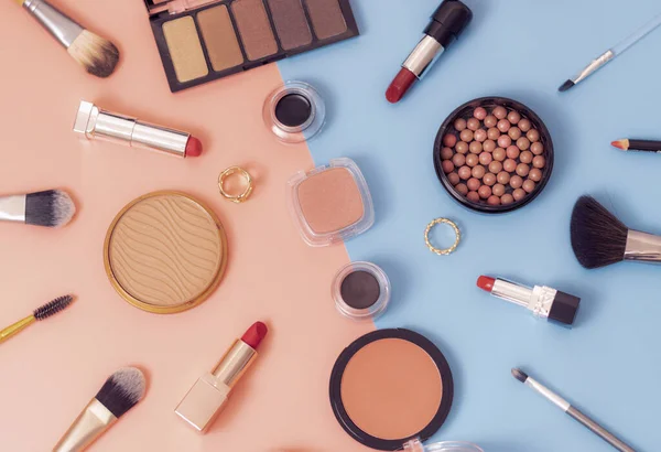 A set of female cosmetics from a face powder box, red lipsticks, makeup brushes, a nude eyeshadow palette, soft eyebrow shadows and rings on a pink-blue background, flat lay close-up. The concept of cosmetics, beauty salon.