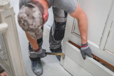 Üniformalı ve gri tekstil eldivenli genç beyaz bir adam, onarımların devam ettiği bir odada dikilirken eski bir ahşap kapıyı söker..