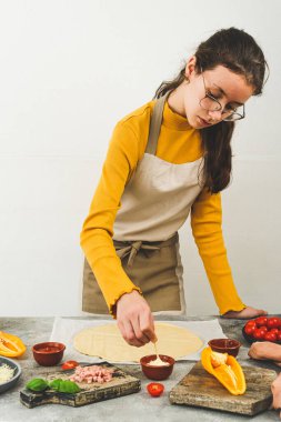 Masada önlüklü, içinde malzemeler ve hamur olan beyaz bir genç kız pizza yapmak için sosu kürdanla karıştırıyor.,