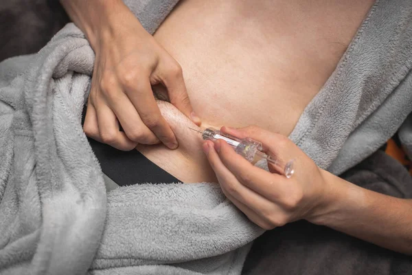 Step 5. Hands of a young caucasian man in a gray robe with a bared belly pinch the skin and hold a syringe with a medicinal liquid for an injection, lying on a bed, close-up side view.Concept step by step instructions for making an injection.