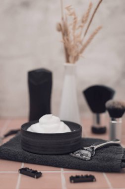Kasede tıraş köpüğü, tıraş fırçası, tıraş fırçası, jilet kafası, makas, tıraş kremi ve vazo, tuğladan yapılma arka planda kurutulmuş çiçek, yakın görüş. Erkek kuaför, güzellik salonu, sakal tıraşı konsepti.