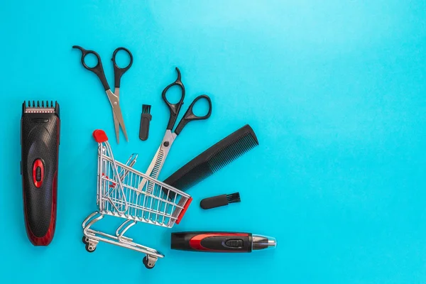 stock image Men's haircut machine, scissors and comb lie in a mini cart on the left on a blue background with copy space on the right, flat lay close-up. The concept of hairdressing and shopping.