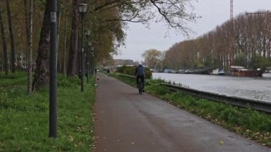 Bir bisikletçi arkadan mavi ceketli ve beyaz şapkalı bir bisikletçi, kanalın bir tarafındaki set boyunca, diğer tarafındaki park boyunca ilerliyor. Yavaş çekimde genişletilmiş bir planın yan görüntüsü..