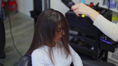 Güzel, beyaz, sarışın kadın kuaför saç kurutma makinesiyle uzun kahverengi saç stili ve fırça fırçası ile kuaförde oturan bir kadın müşteriye, yakın plan ve yavaş çekim..