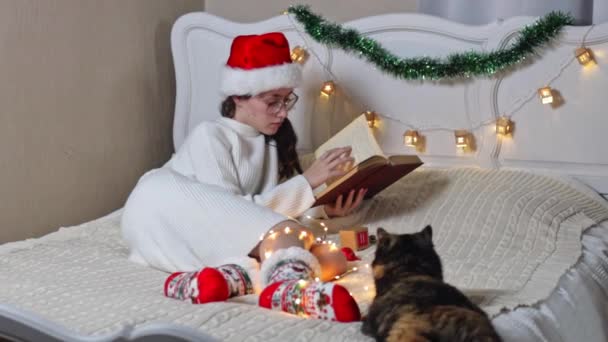 戴着眼镜的美丽的高加索少女 戴着圣塔克洛斯帽 阅读一本书 翻阅躺在床上的一页 旁边放着一盒来历 棒棒糖和一只猫 — 图库视频影像