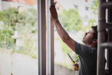 Kıvırcık kahverengi saçlı yakışıklı beyaz bir adam odanın içinde kağıt peçeteyle pencere çerçevesini silerken bir yandan yenileniyor, bir yandan da seçici bir odak noktasıyla yan plan çekiliyor..