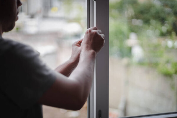 Handsome caucasian young brunette man with curly hair cleans the inner opening of the window frame with his hands, close-up side view with selective focus. The concept of home renovation, washing window frames.