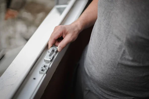 Young caucasian man fitting metal fittings for a plastic window frame in a room where renovations are taking place, close-up side view with selective focus. The concept of home renovation, washing window frames.