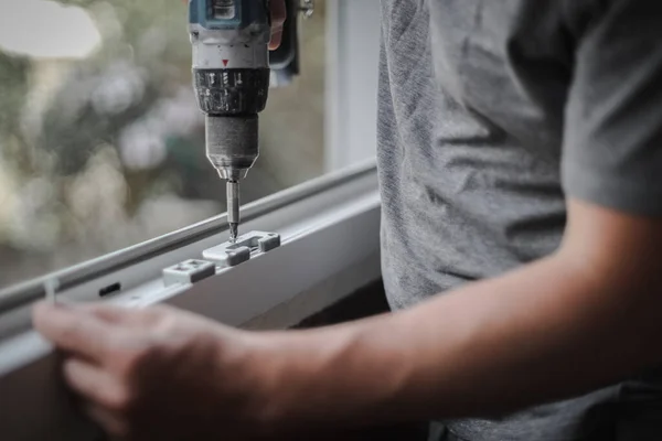 Young caucasian man installs metal fittings for plastic window frame with a drill, tightening a screw in a room where renovations are taking place, close-up side view with selective focus. The concept of home renovation, washing window frames.