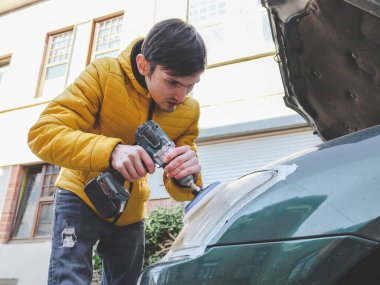 Sarı ceketli genç beyaz bir adam mikrofiber diskle matkap tutuyor ve şehir caddesinde otururken arabasının ön farını parlatıyor, aşağıdan yakın çekim görüntüsü. Konsept: