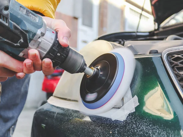 The hands of a young caucasian man are polishing the headlights of his car with a washing soap with a drill and a sponge disc, close-up side view. The concept of headlight polishing, car washing.