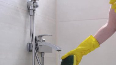 Beyaz tenli, sarı eldivenli genç bir kadının elleri musluğun altına sünger ıslatıyor. Sonra da temizlemeye başlamak için parmaklarıyla banyoyu kapatıyor. Yan görüntüyü yavaş çekimde çekiyor. Tuvalet konseptini temizlemek..