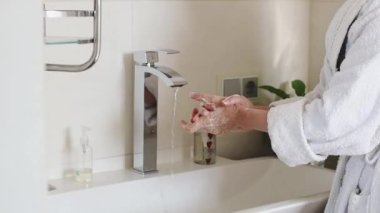 Beyaz bornozlu beyaz bir kadın ellerini sabunla yıkıyor ve banyodaki musluktan akan temiz su akıntısının altında ağır çekimde yan görüş açısını kapatıyor. Konsept vücut bakımı, sağlıklı yaşam tarzı.