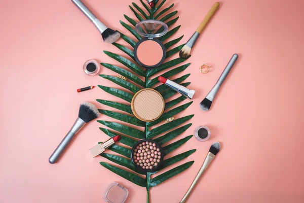 A set of cosmetics from face powder boxes, makeup brushes, red lipstick, eye shadow and rings on a palm tree branch in the center on a pink background, flat lay close-up of sharpness. The concept of cosmetics, beauty salon.