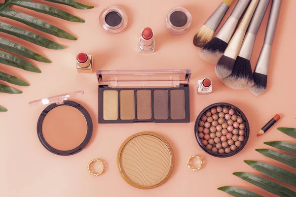 A set of cosmetics from a palette of nude eye shadows, face powder, makeup brushes, rings and palm branches on a pink background, flat lay close-up of sharpness. The concept of cosmetics, beauty salon.